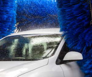 A white car is passing through an automated car wash with blue brushes and water spraying, ensuring routine car wash practices that contribute to vehicle longevity.