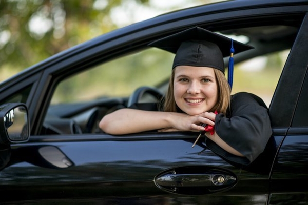 A smiling young woman in graduation attire leaning out of a car window, holding a diploma and car wash packages.