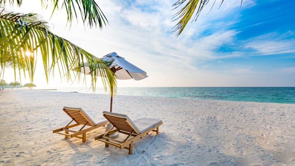 Two wooden lounge chairs with white cushions and a white umbrella are set up on a sandy beach, overlooking a calm, blue ocean under a partly cloudy sky. Perfect for some summer travel relaxation, palm fronds frame the serene scene.