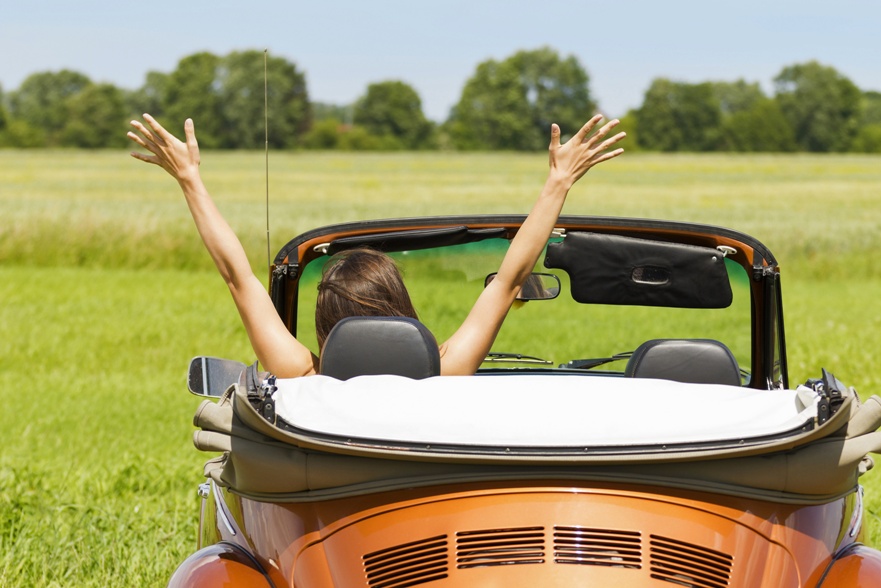 Person in a convertible car with arms raised, soaking in the joy of a summer road trip, facing a field with green grass and trees in the background on a clear day.